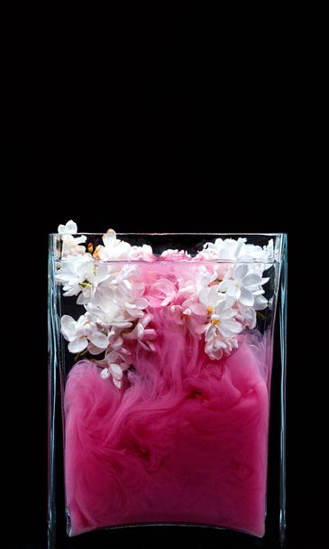 Lila kukkia ja mustetta pyörii vedessä lasimaljakko mustalla pohjalla
 - Valokuva, kuva