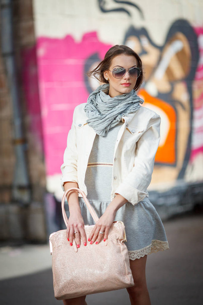 Güzel esmer genç kadın güneş gözlüğü, hafif bir ceket ve sokakta yürüme çanta - Fotoğraf, Görsel