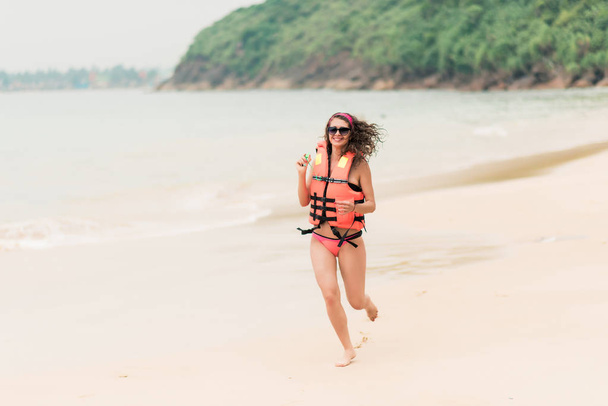 Happy girl running along the beach near the surf in orange life vest - Φωτογραφία, εικόνα