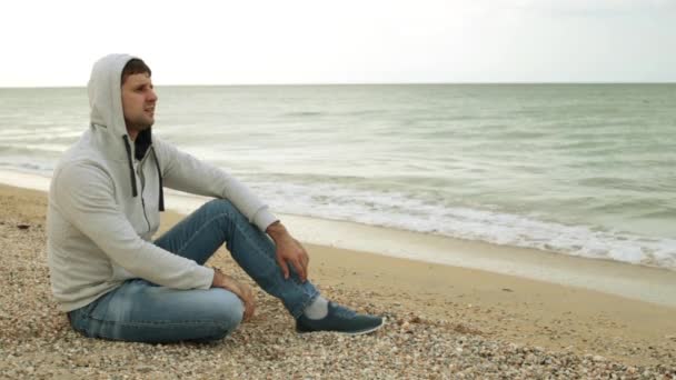 Ένας άνθρωπος αντανακλά στην παραλία. Ένας μοναχικός άνθρωπος για το φόντο της θάλασσας. Ένας άνδρας κάθεται στην παραλία με φόντο κύματα. - Πλάνα, βίντεο