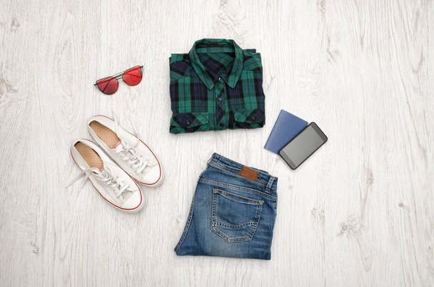 Kék-zöld kockás ing, szemüveg, cipők, farmer, telefon és útlevél. Fából készült háttér. Divatos fogalom, felülnézet - Fotó, kép
