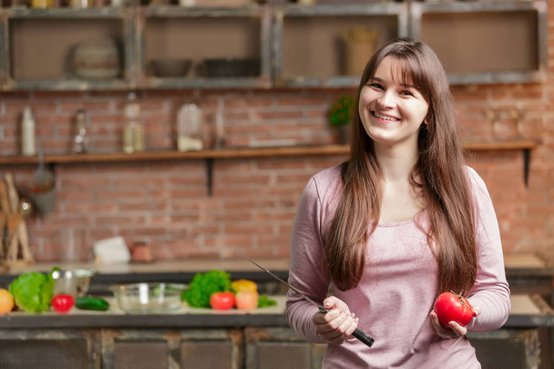 woman is standing in the kitchen near the table with fresh vegetables. girl is holding a knife and a tomato in her hands - Fotografie, Obrázek