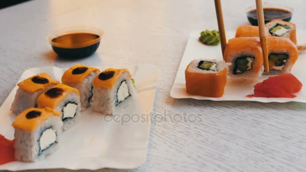 Delicioso sushi con salmón, queso, wasabi, jengibre rosa, salsa de soja que se toman con palos de madera sobre un elegante fondo de madera blanca
 - Metraje, vídeo