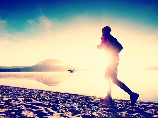 Silhouette des sportlich aktiven Mannes, der bei Sonnenaufgang am Strand des Sees läuft. Gesunder Lebensstil.  - Foto, Bild