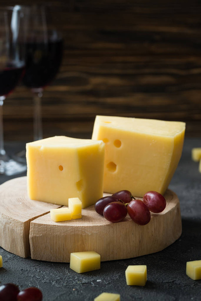 Swedish hard yellow cheese with holes chopped with red grapes on wooden slices and glasses with red wine - Fotó, kép