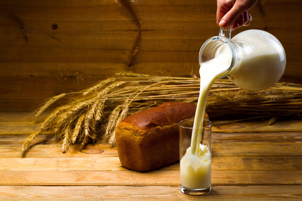 Milk from a glass jug poured into a glass, a loaf of rye bread, ears of corn - Zdjęcie, obraz
