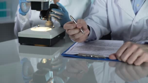 Lab worker filling paper forms, his assistant viewing samples under microscope - Footage, Video