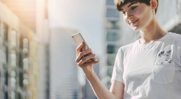 Hipster-Mädchen trägt weißes T-Shirt und tippt auf Touchscreen des Handys, Frau genießt Abendspaziergang und benutzt ihr Smartphone - Foto, Bild