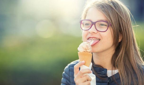 Girl. Teen. Pre teen. Girl with ice cream. Girl with glasses. Girl with teeth braces. Young cute caucasian blond girl wearing teeth braces and glasses. Portrait of a smiling young girl - 写真・画像