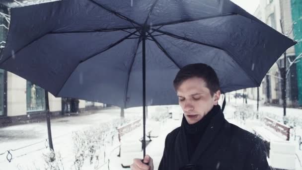 Young man in coat with umbrella under snowfall using smartphone on alley - Imágenes, Vídeo