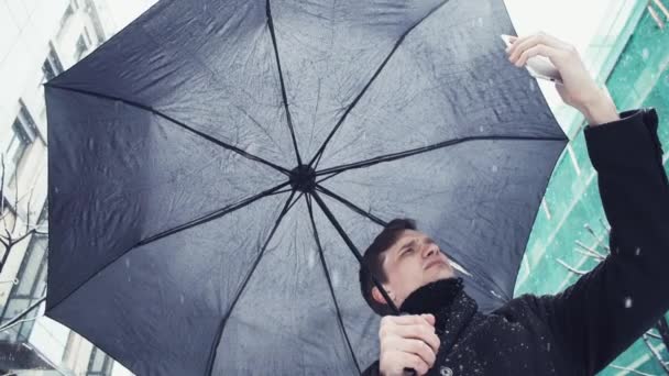 Young man with umbrella under snowfall making selfie photo using smartphone - Metraje, vídeo