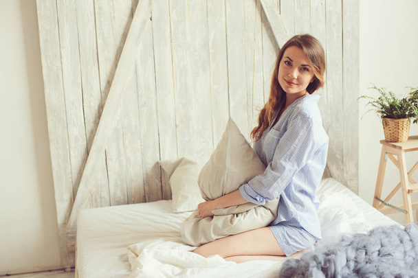 young woman in pajama wake up in the morning in cozy scandinavian bedroom and sitting on bed with white bedlinen. Casual lifestyle in modern interior - Foto, Imagen