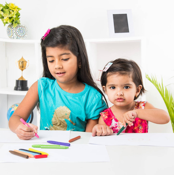 két imádnivaló indiai kislányok nővérek forgalmas a színe vagy rajz vagy festmény, pasztell színek használatával vagy vázlat, toll, otthon az asztalnál - Fotó, kép
