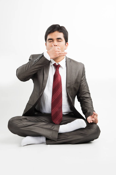 front view of an indian young businessman meditating or practicing yoga or pranayama or breathing exercise in corporate attire in the office or isolated over white background - Photo, Image