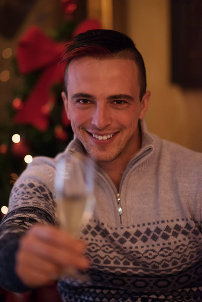 Portrait of a happy young man with a glass of champagne celebrating winter holidays at home beautifully decorated for Christmas - Fotó, kép
