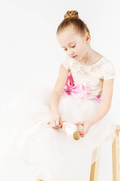 A small ballerina dresses or takes off her pointe isolated on white background. Slender little ballerina girl in white dress and in pointe shoes - Fotoğraf, Görsel