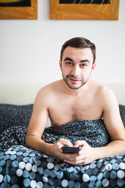 attraktiver junger Mann mit Bart mit Smartphone und SMS, während er auf seinem Bett liegt - Foto, Bild