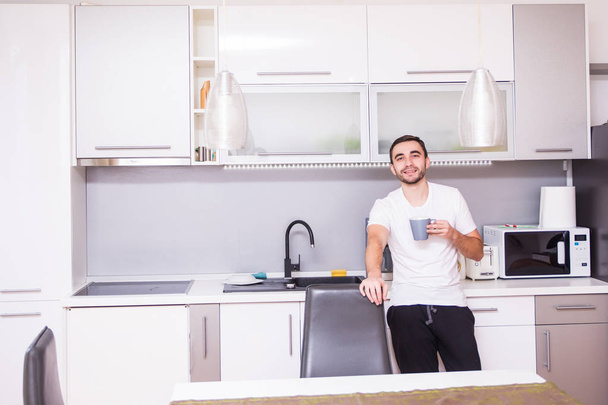 Portrait of man standing in kitchen holding a cup of coffee looking away in thought. Relaxed young man having coffee at home in kitchen - Foto, Imagen