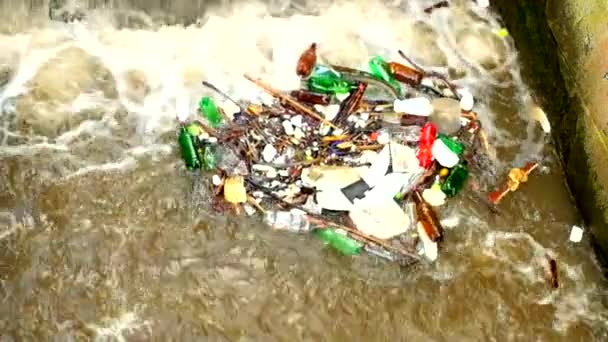 Slow-motion. Plastic flessen en andere rommel draaien op de waterstand. Draaien van vuil water boven weir op riviertje met modderig water. Stenen muur van weir  - Video