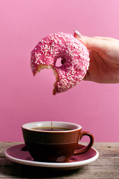 Woman hand holding colorful donut with sprinkles on a wooden table and pink background. Cup of coffee. Concept of food. Donut with bite missing Close up. Pastel color  - Foto, imagen