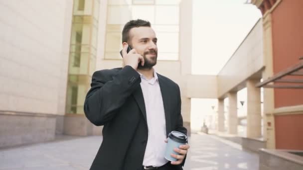 Dolly shot of young bearded business man talking at smartphone with cup of coffee near modern office buildings - Imágenes, Vídeo