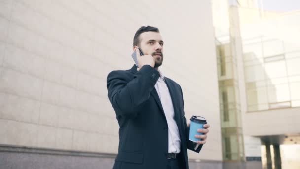 Lage hoek schot van jonge bebaarde zakenman te praten op smartphone met kop koffie in de buurt van moderne kantoorgebouwen - Video