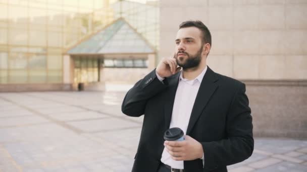 Young bearded business man talking at smartphone with cup of coffee near modern office buildings - Video