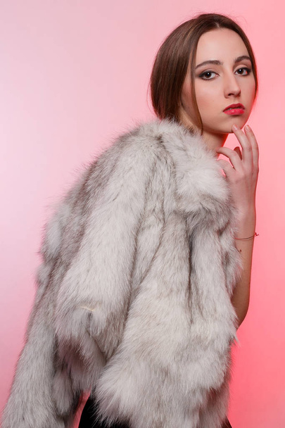 Fashionable beautiful woman in a gray fur coat of artificial fur on a red background stands frowning - Valokuva, kuva