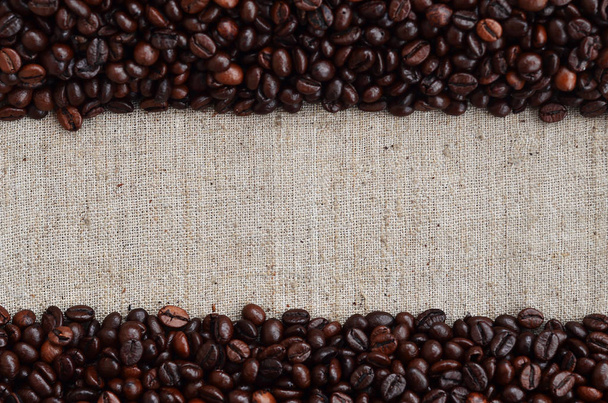 Texture of a gray canvas made of old and coarse burlap, on which lies a certain amount of brown coffee beans. Top view of sackcloth of light gray color under sunlight with pile of coffee beans - Fotografie, Obrázek
