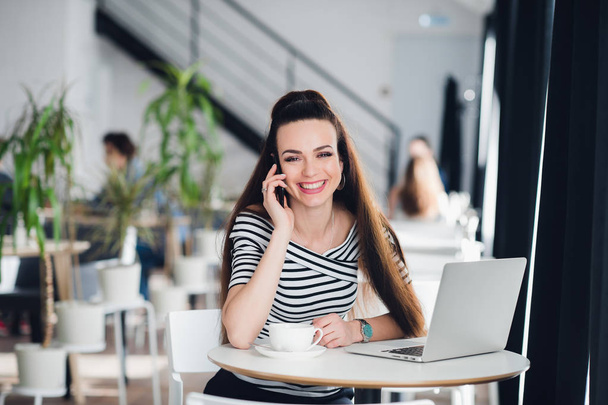 Donna esecutiva che sorride e lavora online con un computer portatile mentre parla al telefono in un caffè
. - Foto, immagini