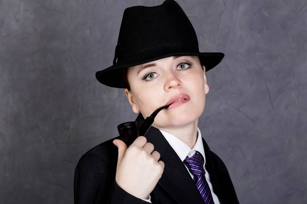 beautiful woman in manly style with smoking pipe on gray background, girl in mans suit and tie, white shirt and hat - Foto, Imagem
