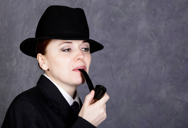 beautiful woman in manly style with smoking pipe on gray background, girl in mans suit and tie, white shirt and hat - Photo, Image