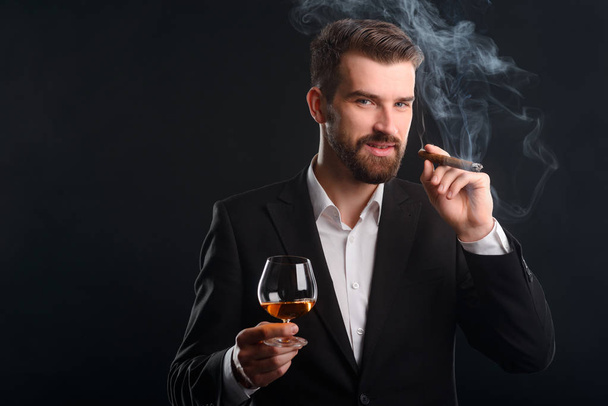 Portrait of successful businessman. The boss is in the dark room holding a glass of premium brandy and smoking fine Cuban cigar. Horizontal shot with black background. - Fotó, kép