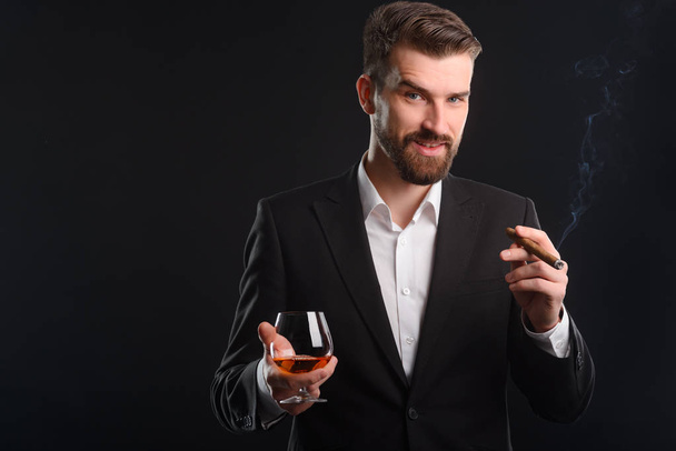 Portrait of solid businessman with black background. Slight smile and uplifted mood of young successful boss. A glass of fine quality brandy and a little fuming cigar in his hands. - Фото, изображение