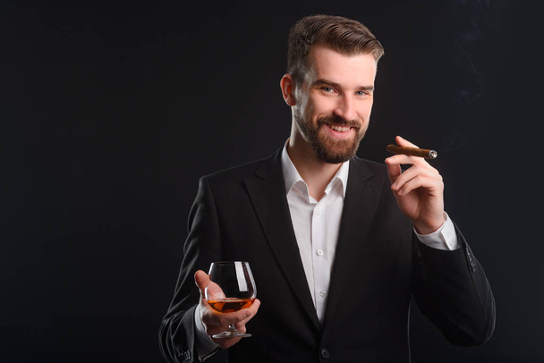 Macho smoking an expensive cigar. Slightly laughing he is holding a glass of brandy and a Cuban cigar in both hands. Dark background with vanishing flows of smoke. - Foto, Bild