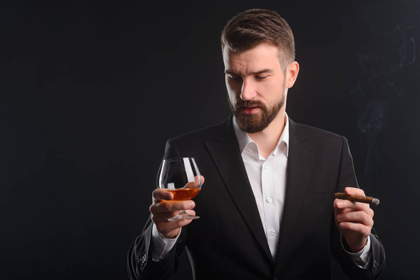 Successful businessman drinking and smoking. Young man having a rest in company with a glass of premium cognac and an expensive Cuban cigar. Studio shot, black background. - Photo, image