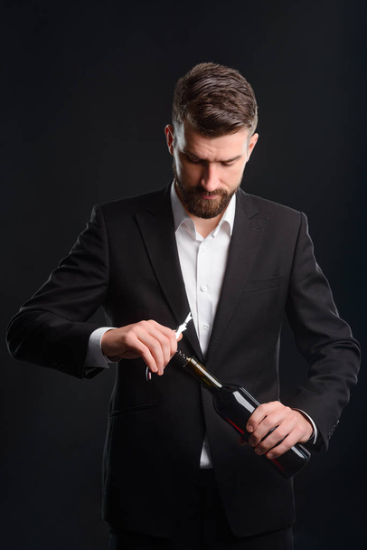 Opening a bottle of red wine. Stylish young man dressed in black suit and white shirt with unfastened top button using a corkscrew. Vertical portrait of sommelier on black background. - Photo, Image