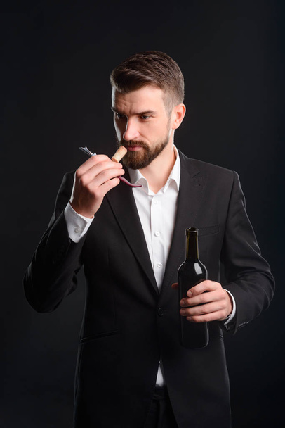 Вино эксперт нюхает пробку, чтобы определить, если вино испорчено. Вертикальный портрет молодого человека в черном костюме и белой рубашке с бутылкой и сомелье-ножом с пробкой в руках. Чёрный фон
. - Фото, изображение