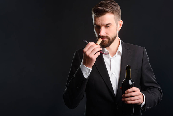 Tasting session of red wine. Young man dressed in formal style clothes has just opened a bottle. Handsome sommelier focused on smelling a cork on black background with empty space to insert your text. - Zdjęcie, obraz