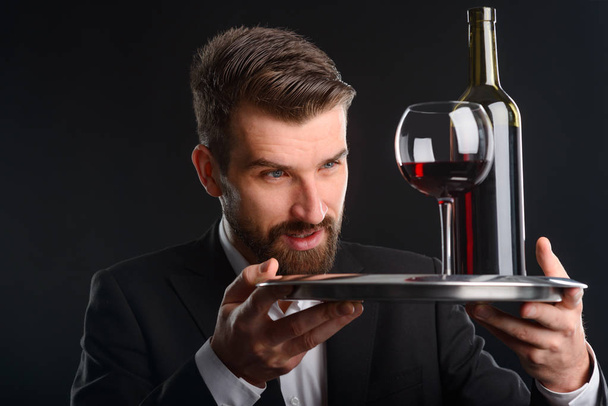 Closeup shot of man with drink. Salver with a glass and a bottle of red wine in both hands of handsome and stylish man standing against black background. - Photo, image