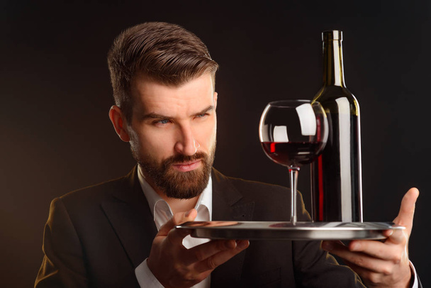 Handsome sommelier judging wine by its appearance. Skillful taster looking narrowly at color and consistence of beverage. A glass and a bottle of red wine on a salver. - 写真・画像