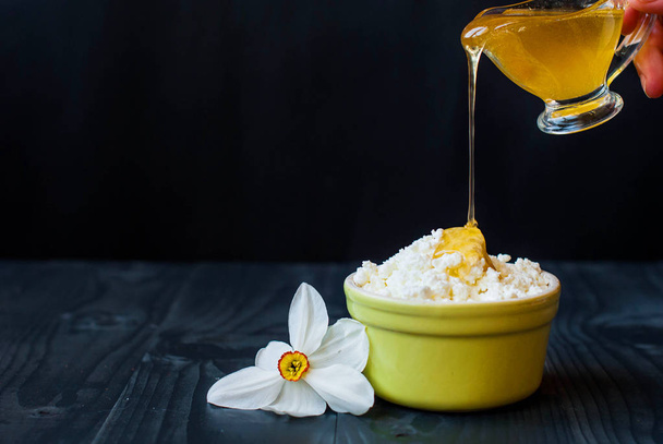 The young cheese is poured with honey. Fresh cottage cheese and honey on a dark background. Hand water honey with cottage cheese. - Photo, Image