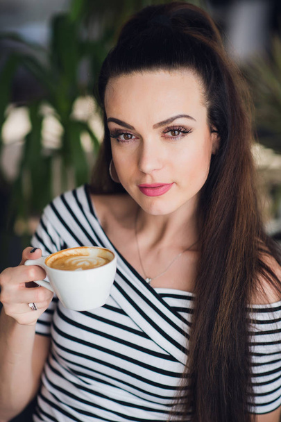 Close up portrait of beautiful but a bit tired woman holding a cup of cappuccino. - Foto, afbeelding