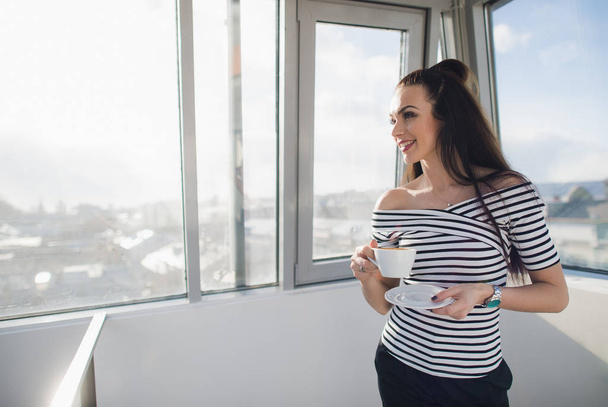 Mujer feliz en blusa rayada sonriendo y de pie en el interior con una taza y mirando a través de la ventana
. - Foto, Imagen