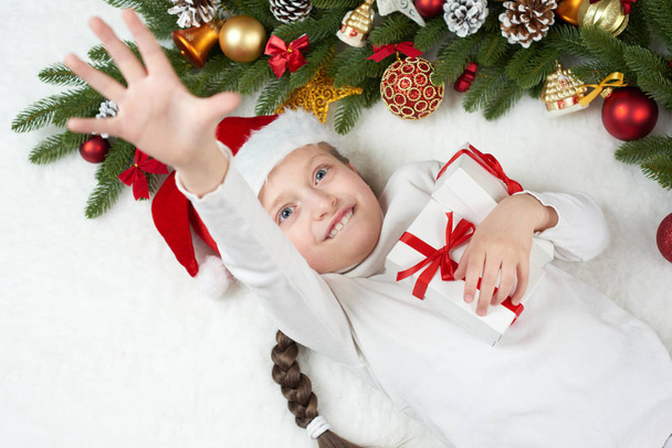 child girl having fun with christmas decoration, face expression and happy emotions, dressed in santa hat, lie on white fur background, winter holiday concept - Photo, Image