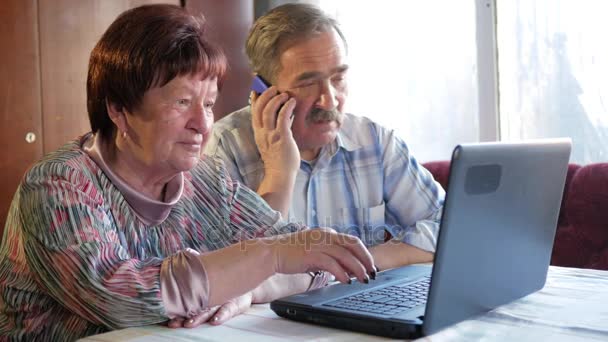 An elderly couple is sitting at home at the laptop. Woman reading news, man talking on mobile phone - Materiał filmowy, wideo