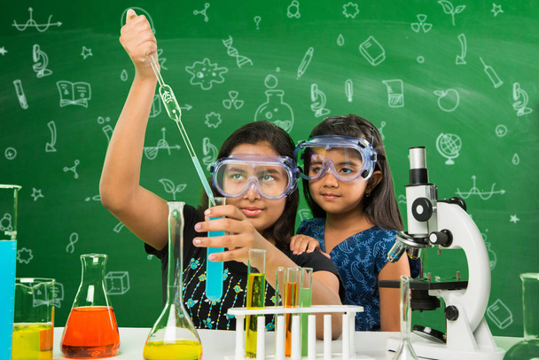 2 cute little indian girls doing science experiment or project in a classroom with green chalkboard having science doodles - Photo, Image