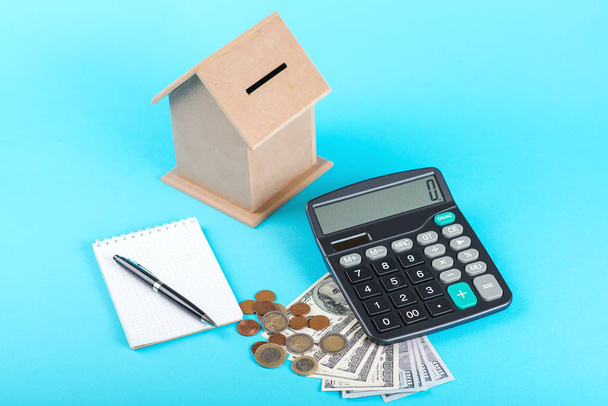 das Konzept der finanziellen Ersparnisse, um ein Haus zu kaufen. Spardose, Dollar, Münzen und Taschenrechner isoliert auf blauem Hintergrund. - Foto, Bild
