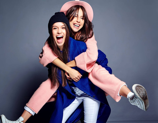 fashion portrait of two smiling brunette women models in summer casual hipster overcoat posing on gray background. Girls holding each other on back  - Photo, Image