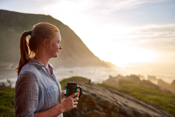Young woman with light ginger hair and a fresh beauty, standing quietly with a flask of coffee in the early morning, looking out at a picturesque coastline - Foto, imagen